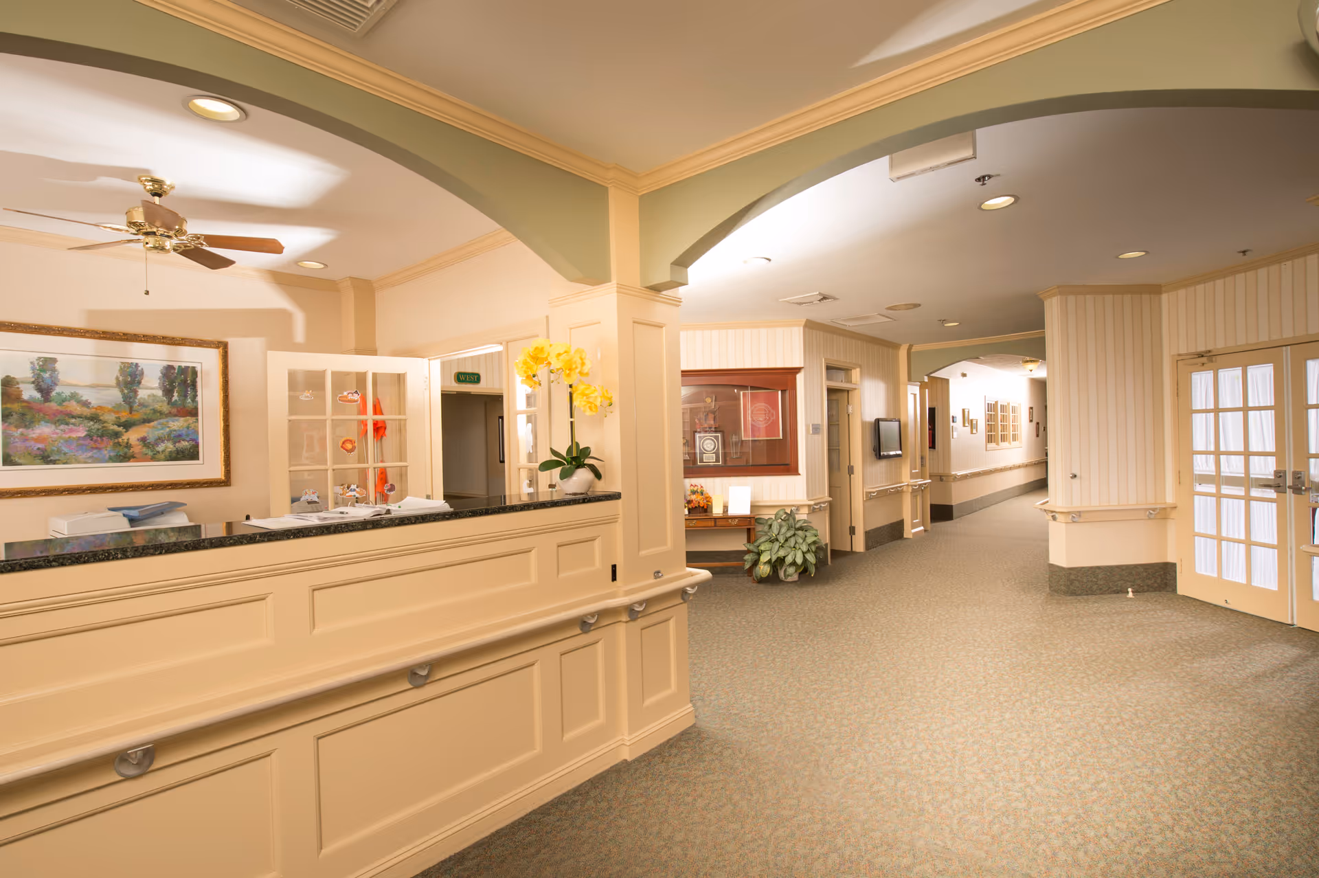 Interior view of a senior living facility hallway with a reception desk on the left, decorated with a yellow orchid and a framed landscape painting on the wall. The hallway has beige walls with white trim, carpeted floors, ceiling lights, and a ceiling fan. There are doors and a display case along the hallway, with handrails on the walls.