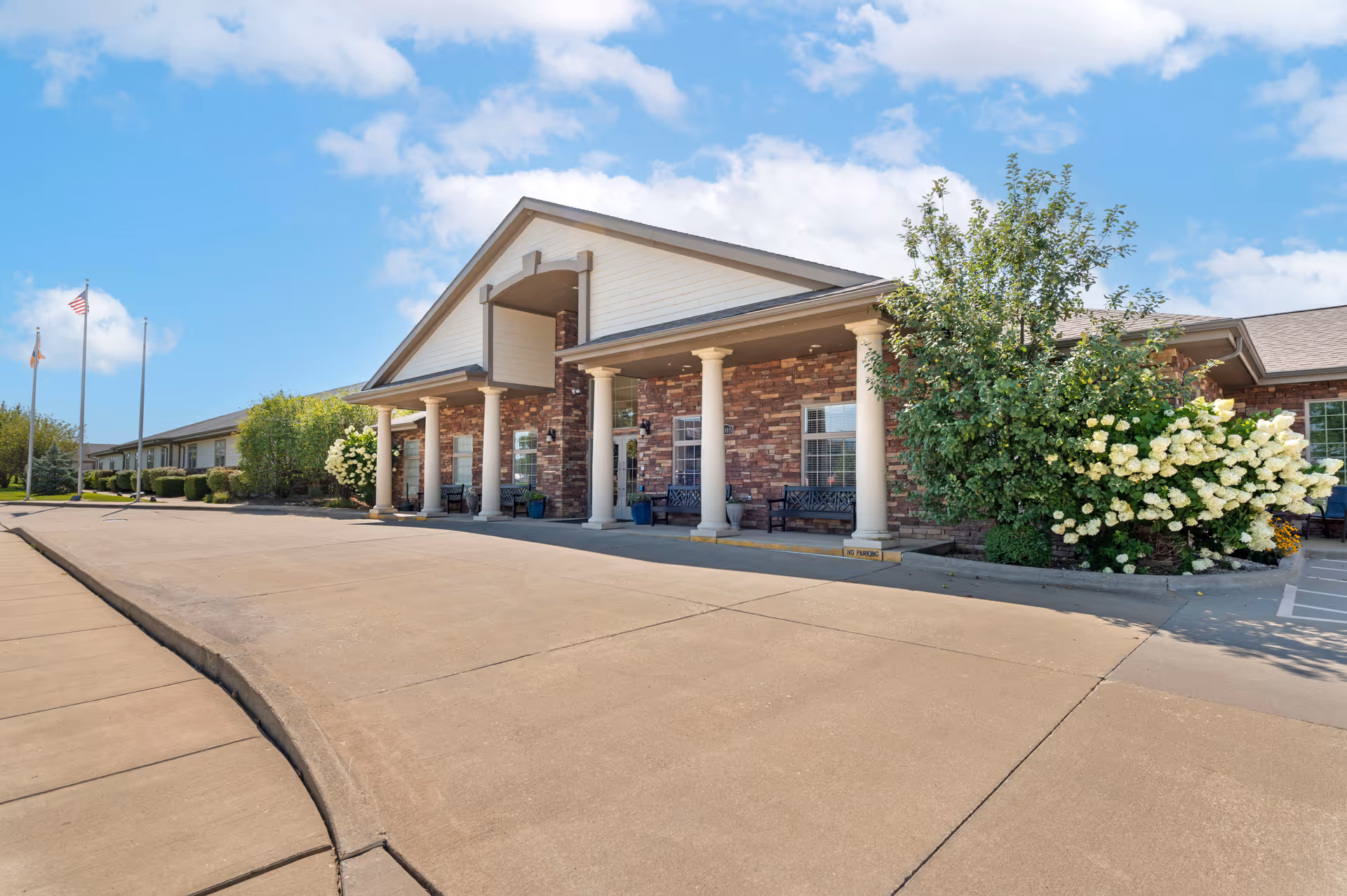 Exterior view of Bridle Brook Assisted Living & Memory Care Community building with a covered entrance supported by white columns, brick facade, benches, and landscaped bushes and flowers under a blue sky with some clouds.