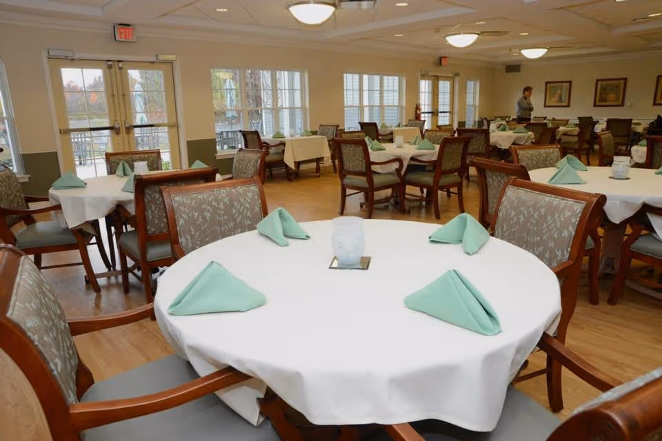 A bright assisted-living dining room with round tables covered in white tablecloths and neatly folded green napkins.