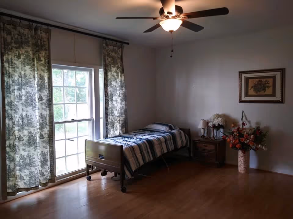 A simple bedroom with a single bed covered in a striped blanket, positioned next to a large window with patterned curtains. There is a wooden nightstand with a lamp and decorative flowers, a framed floral picture on the wall, and a ceiling fan with a light fixture above. The floor is wooden.