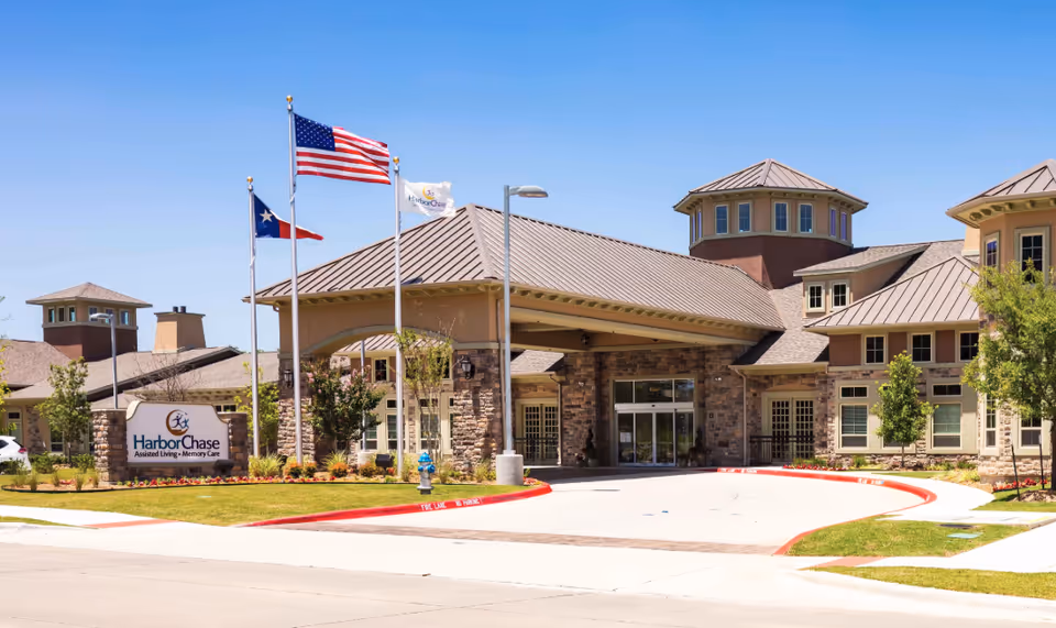 Exterior view of Harbor Chase assisted living and memory care facility with a covered entrance, stone and beige walls, and three flagpoles flying the American, Texas, and Harbor Chase flags under a clear blue sky.