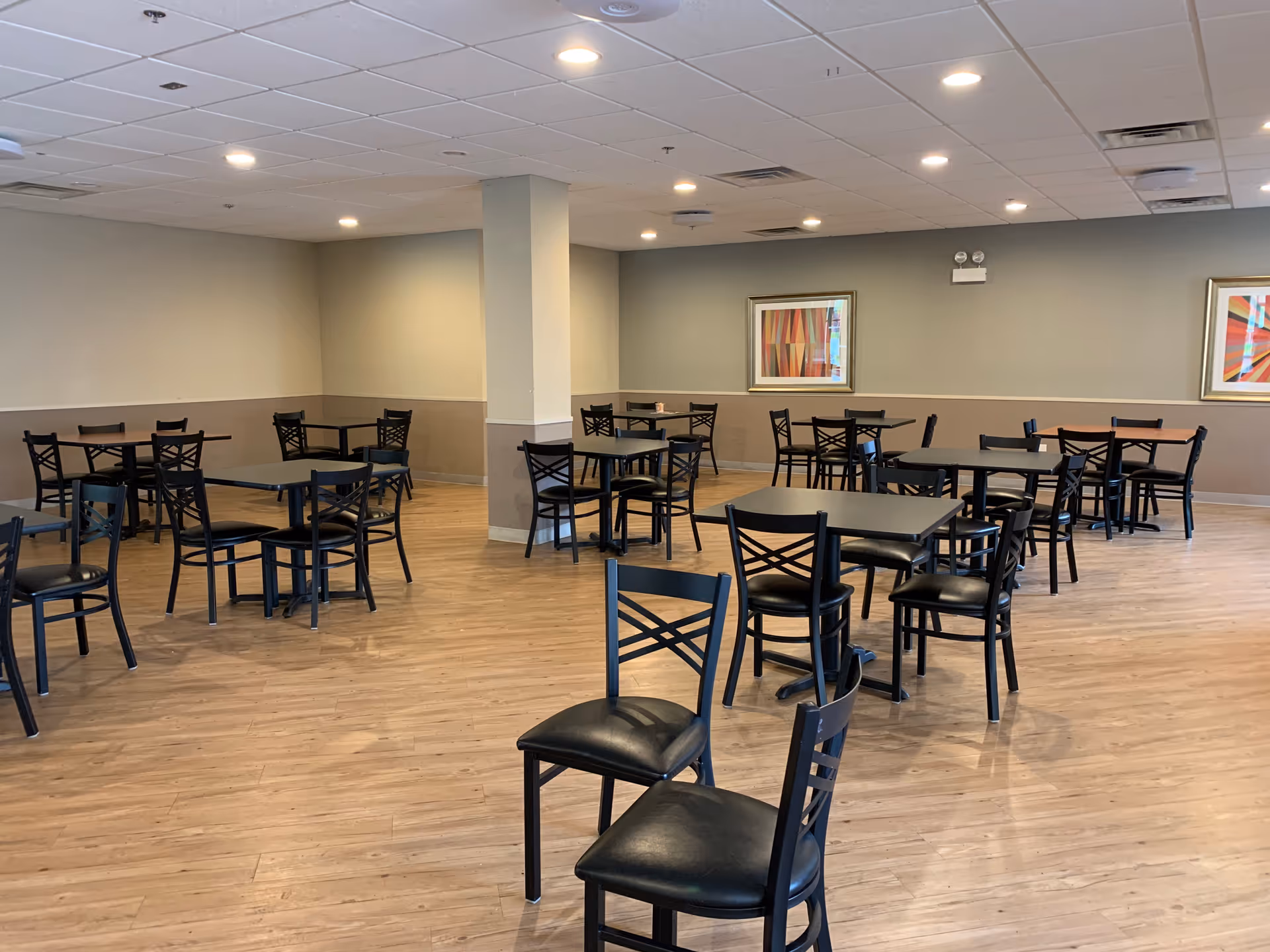 A spacious dining room with multiple black tables and chairs arranged on a wooden floor. The walls are painted in neutral tones with two colorful abstract paintings hanging. The ceiling has recessed lighting and a white drop ceiling.