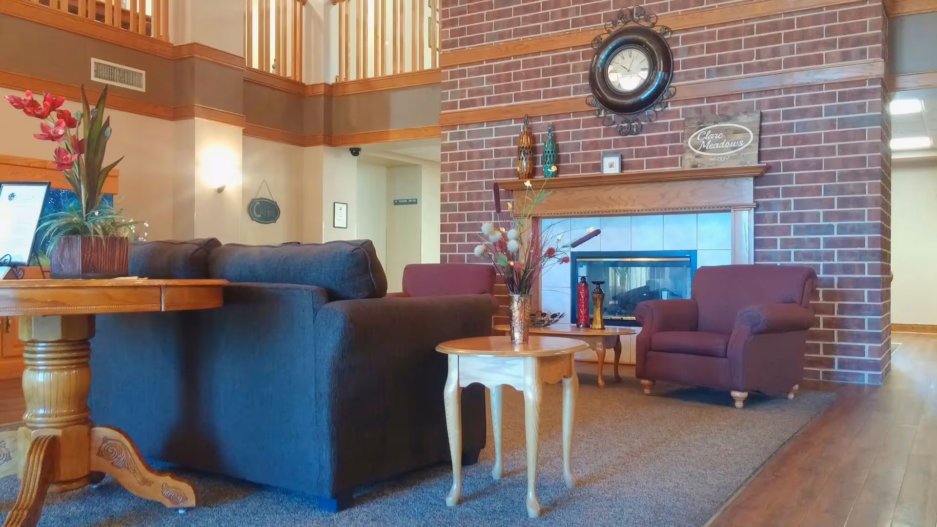 A cozy living room area with a dark gray sofa, a wooden side table with a vase of flowers, two maroon armchairs, and a brick fireplace with decorative items and a clock above it. The room has wooden flooring and a high ceiling with a balcony railing visible above.