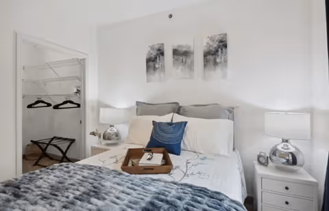 A neatly made bed with white and gray bedding, a blue decorative pillow, and a wooden tray on top. There are two bedside tables with matching lamps on each side of the bed. Above the bed, three black and white abstract art pieces are hung on the white wall. To the left, an open closet with hangers and a luggage rack is visible.