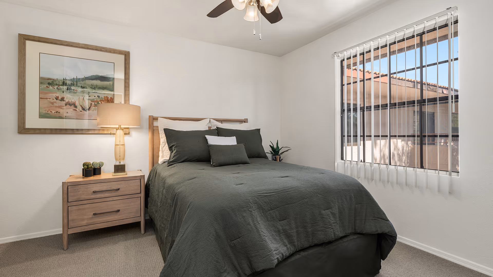 A bedroom with a neatly made bed featuring dark green bedding and multiple pillows. There is a wooden nightstand with two drawers beside the bed, holding a lamp and small potted plants. A framed landscape painting hangs on the wall above the nightstand. A window with vertical blinds lets in natural light, and a ceiling fan with lights is visible above.