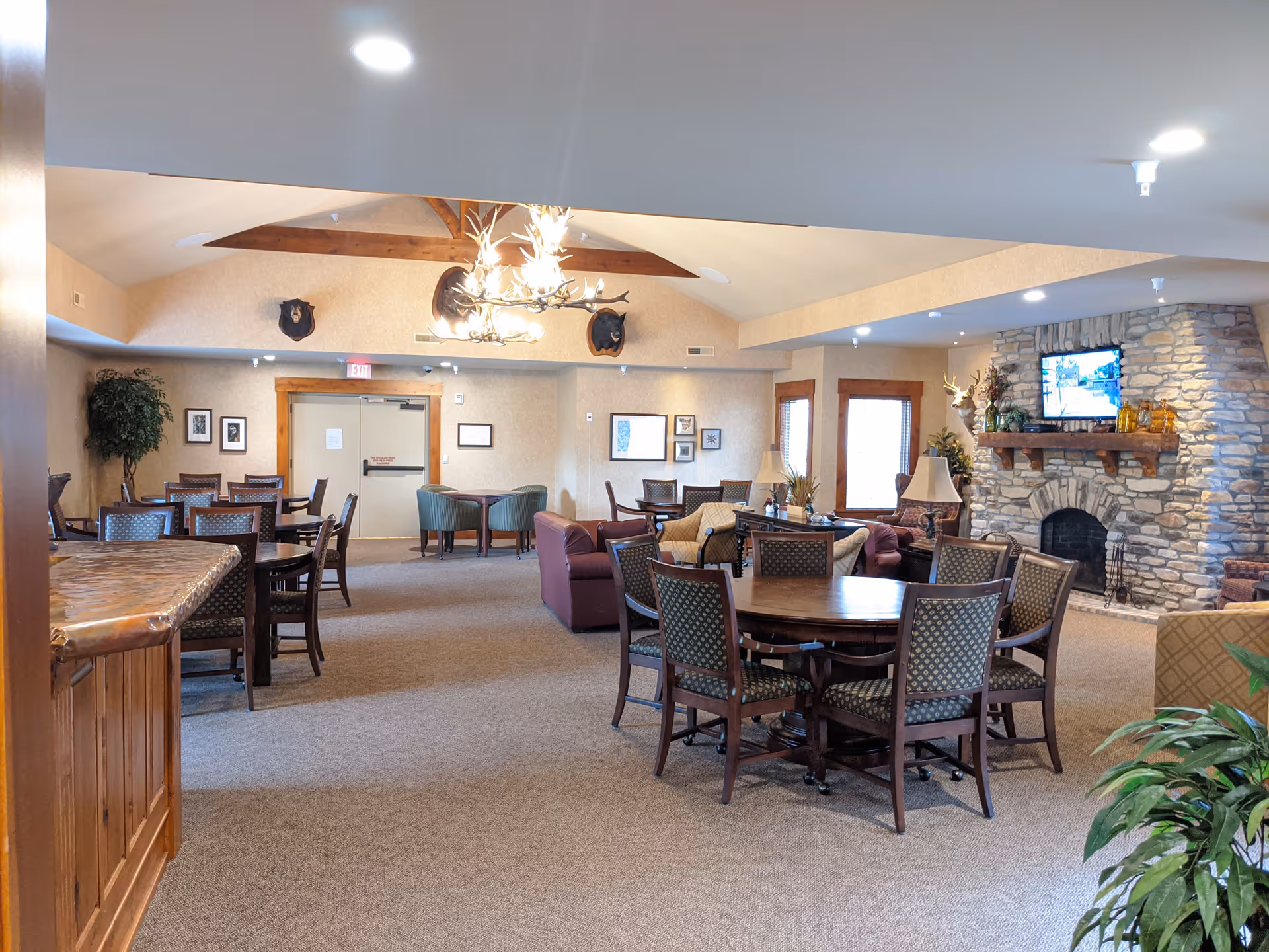 A spacious common area in a senior living facility featuring multiple round tables with chairs, a stone fireplace with a mounted TV above it, comfortable sofas, armchairs, and decorative elements such as plants and framed pictures on the walls. The ceiling has wooden beams and a chandelier with antler design.