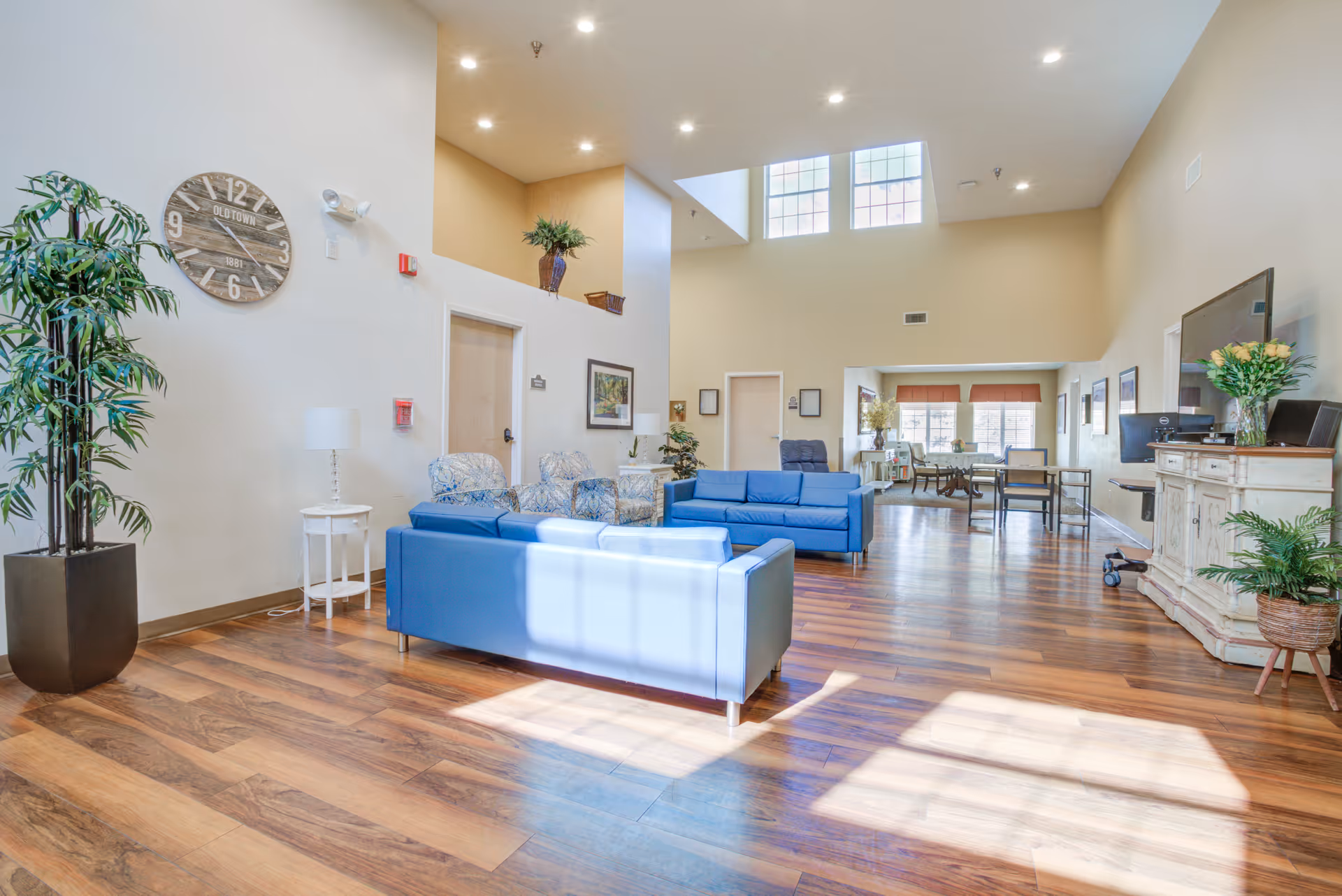 A bright and spacious senior living facility common area with wooden flooring, blue sofas, patterned armchairs, a large wall clock, potted plants, and a TV on a white cabinet. Large windows allow natural light to fill the room.