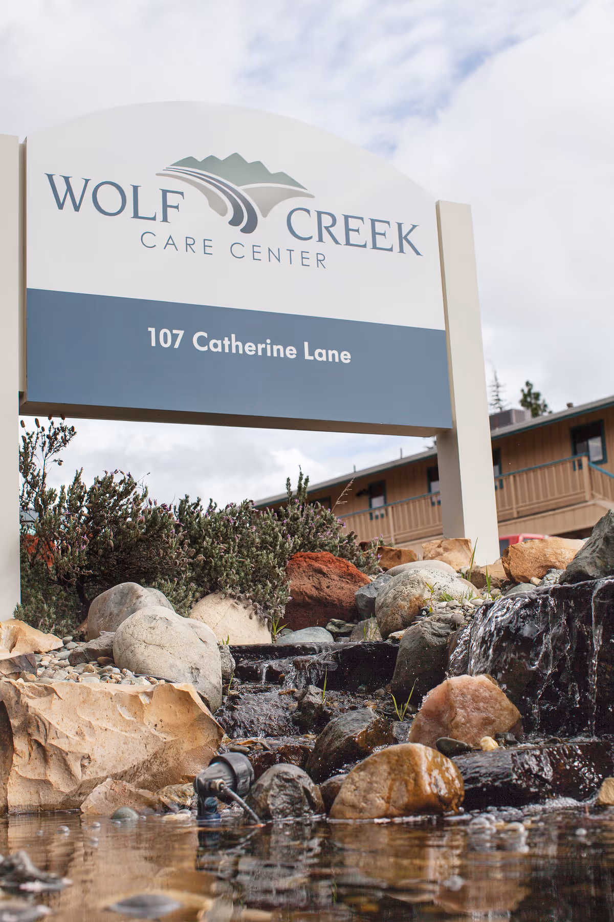 Sign reading 'Wolf Creek Care Center' and '107 Catherine Lane' above a rock-lined pond and water feature in front of the building.