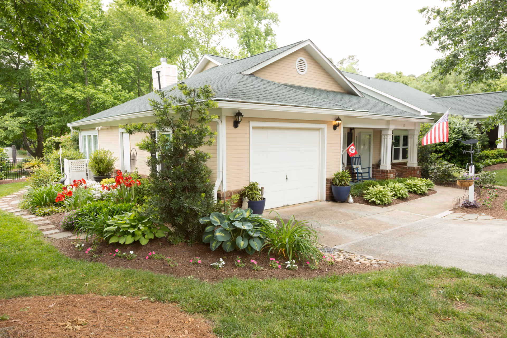 Front exterior of a single-story beige building with a garage, porch, American flag, and landscaped garden.