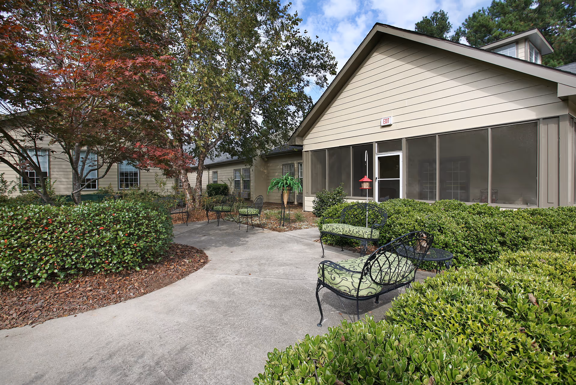 Outdoor patio area at The Carriages of Mt. Zion featuring metal chairs with green cushions, a small round table, and surrounding greenery including bushes and trees with some red leaves. The building exterior is beige with a screened porch and an exit door.
