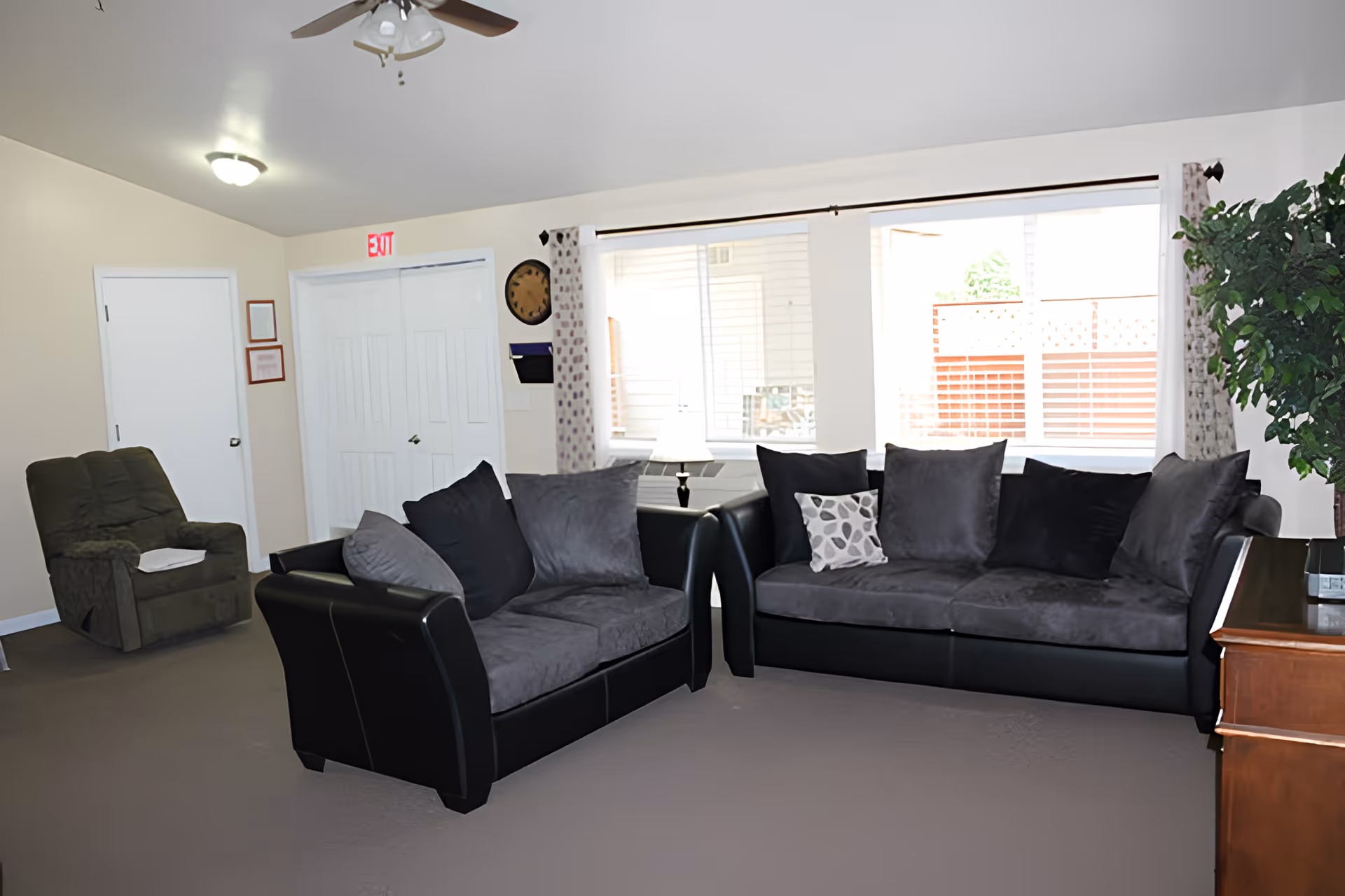 A living room area with two black and gray sofas arranged around a wooden cabinet. There is a green recliner chair with a white cloth on it in the corner. The room has beige walls, a ceiling fan with lights, a large window with white curtains, and a potted plant on the right side. An exit sign is visible above a set of white double doors.