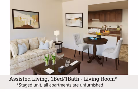 A staged living room in an assisted living apartment featuring a beige patterned sofa with light blue pillows, a dark wooden coffee table with decorative items, a round dining table with three upholstered chairs, and a kitchen area with wooden cabinets and a stove in the background. Two framed abstract paintings hang on the walls.
