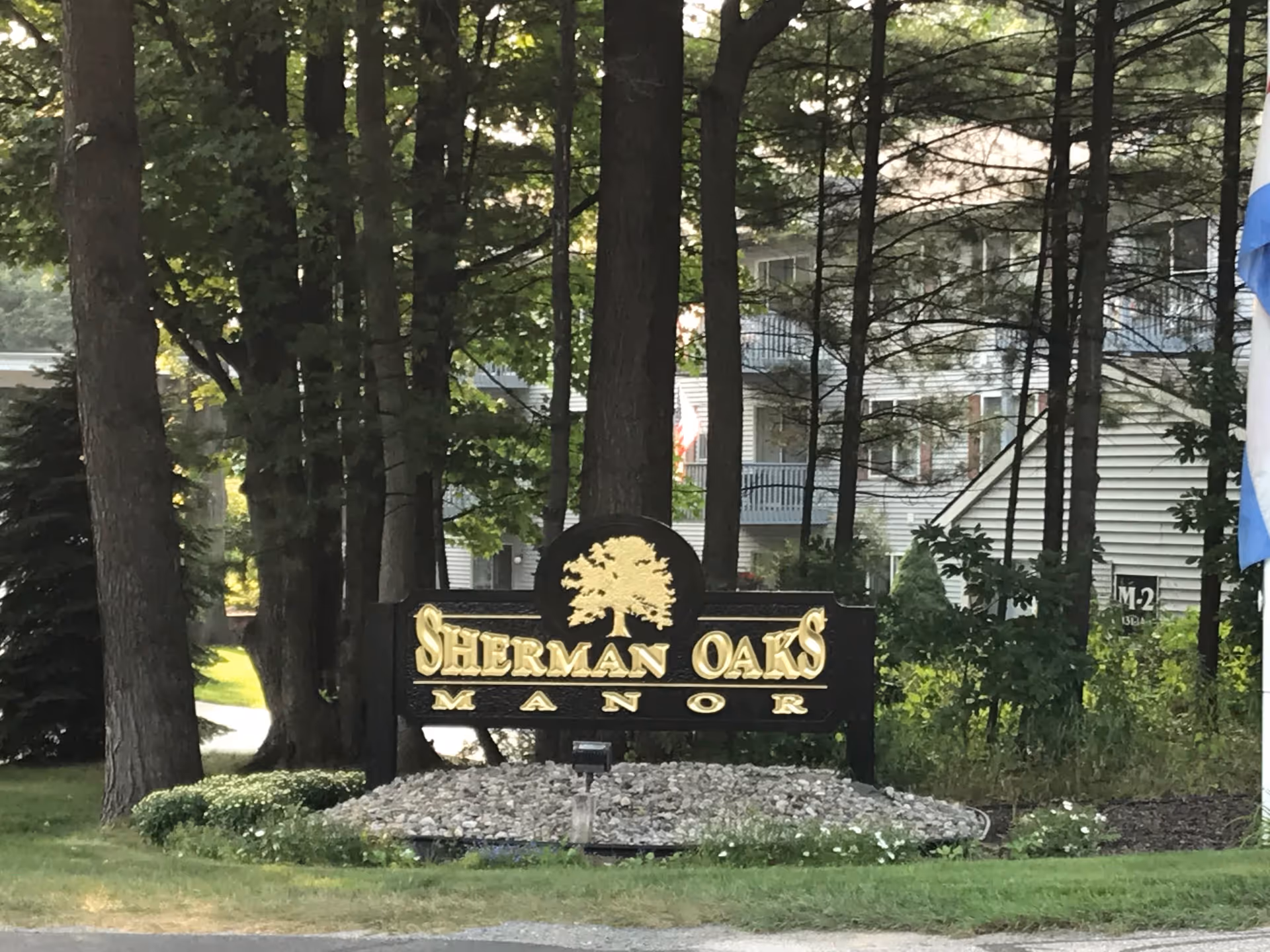 Wooden entrance sign reading 'Sherman Oaks Manor' set among trees and landscaping with the facility building visible behind.