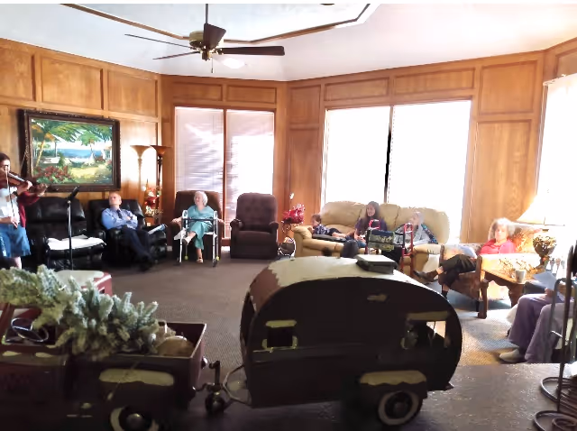 A wood-paneled communal living room with seniors seated around sofas and chairs and a small toy camper in the foreground.