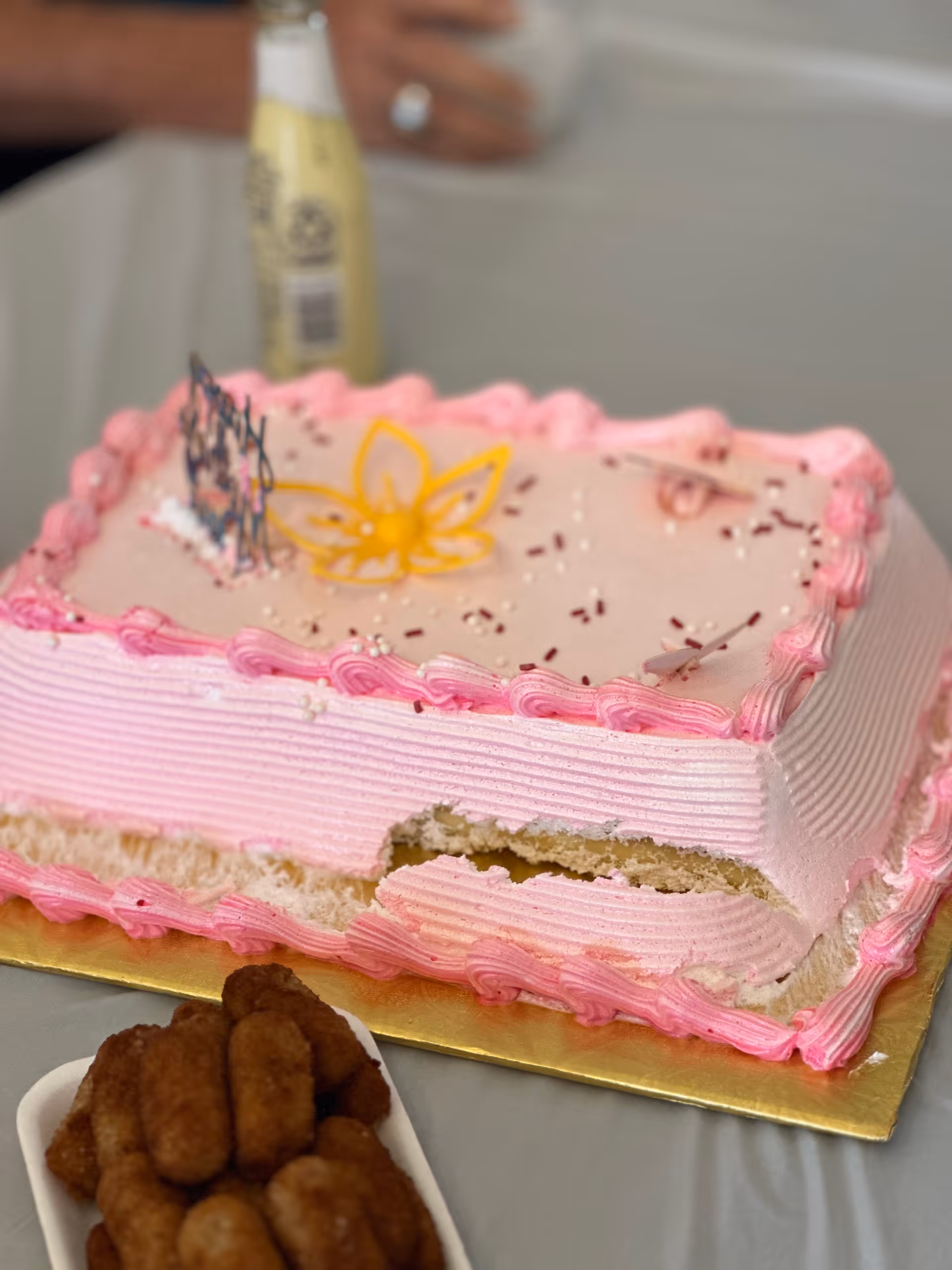 Partially eaten rectangular pink frosted sheet cake with a yellow flower decoration on a table and a small plate of fried dough bites.