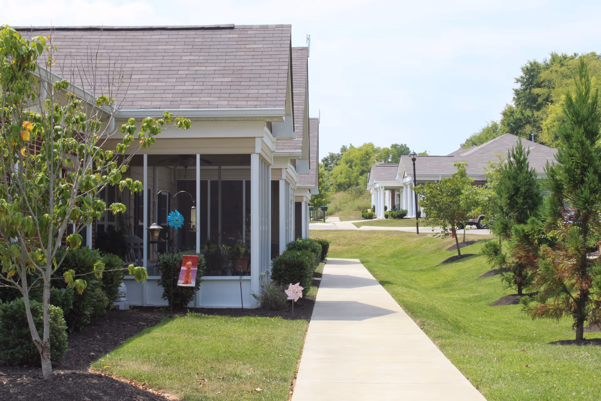 A paved walkway runs alongside a row of single-story buildings with screened porches, surrounded by well-maintained green lawns, small trees, and shrubs under a clear sky.