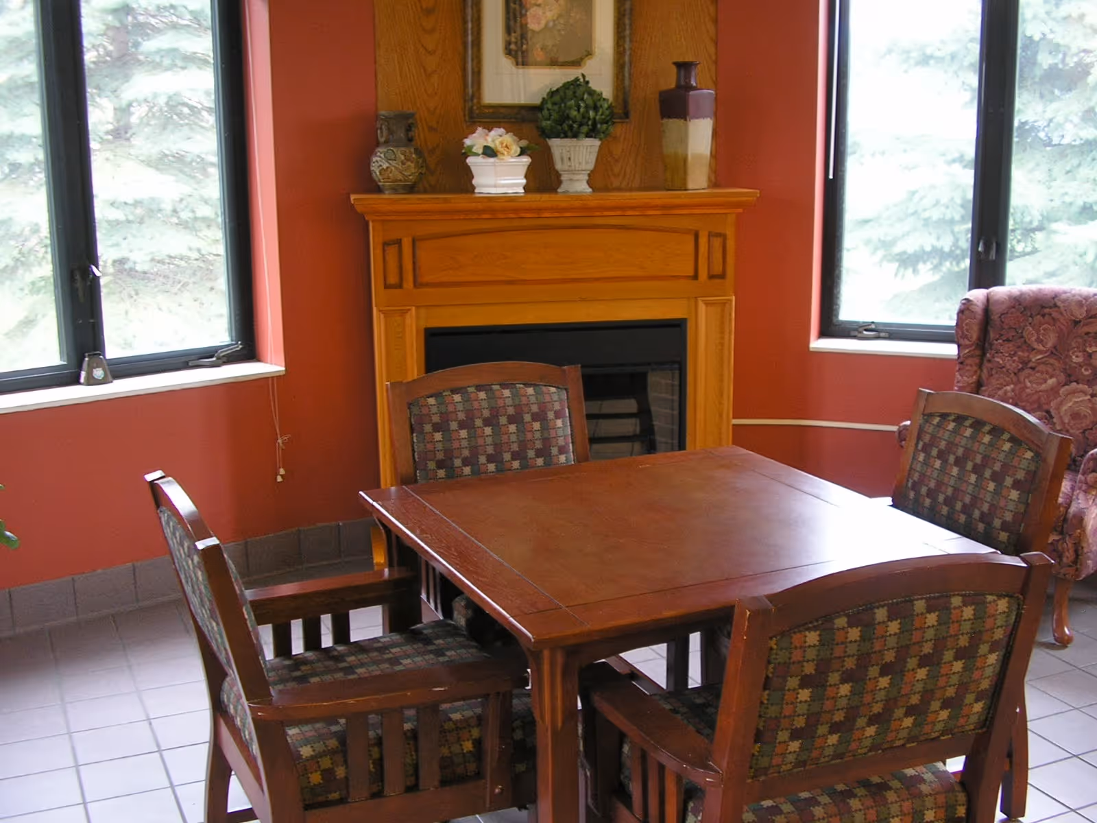 A wooden square dining table surrounded by four upholstered chairs sits in front of a wooden fireplace between two windows in a cozy room.