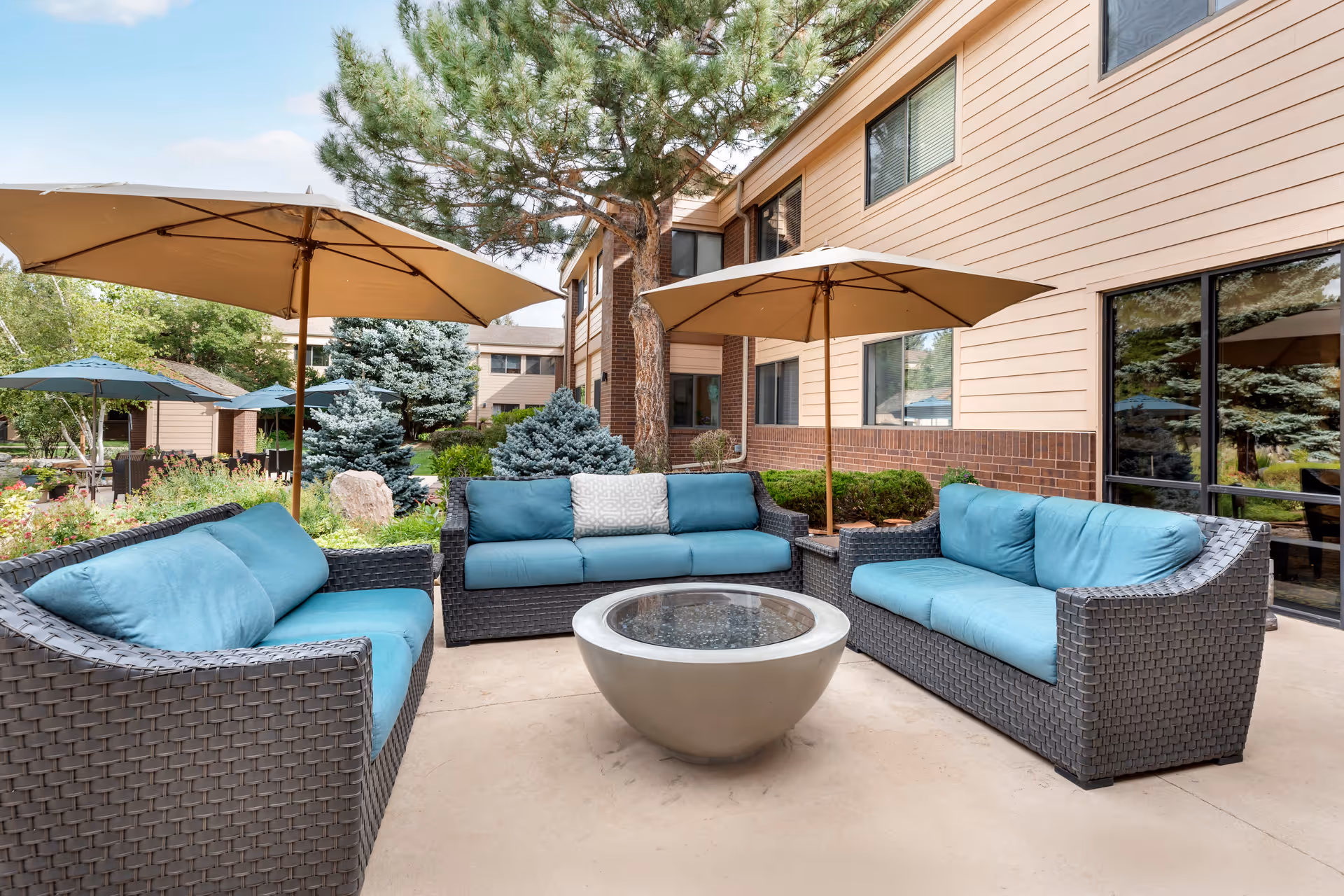 Outdoor patio area with three wicker sofas featuring blue cushions arranged around a circular fire pit table. Two large beige umbrellas provide shade. The patio is adjacent to a beige building with large windows and surrounded by greenery including trees and shrubs.