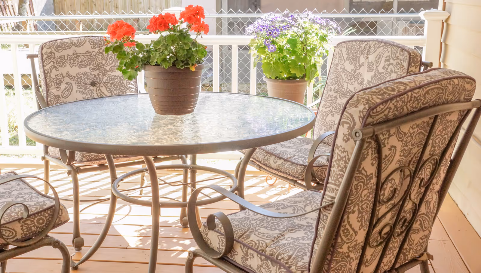 A round glass-top patio table with four cushioned metal chairs featuring a paisley pattern. Two potted plants with red and purple flowers are placed on the table and in the background. The setting is on a wooden deck with a white railing.
