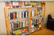 Two wooden bookshelves filled with books and board games, placed against a beige wall. To the right, part of a black armchair with a striped blanket draped over the arm is visible.