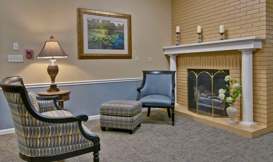 A cozy living room area with two patterned armchairs, a matching ottoman, a side table with a lamp, a framed painting of a pond with lily pads on the wall, and a brick fireplace with a white mantel decorated with three candles and a vase of flowers.