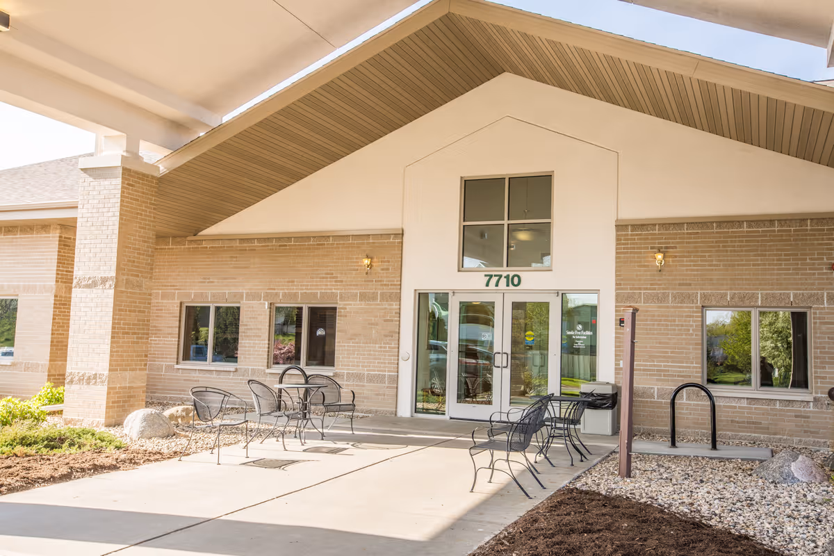 Entrance of Coventry Village facility showing a covered walkway with metal chairs and tables outside. The building has a brick exterior with large glass double doors and the number 7710 above the entrance.
