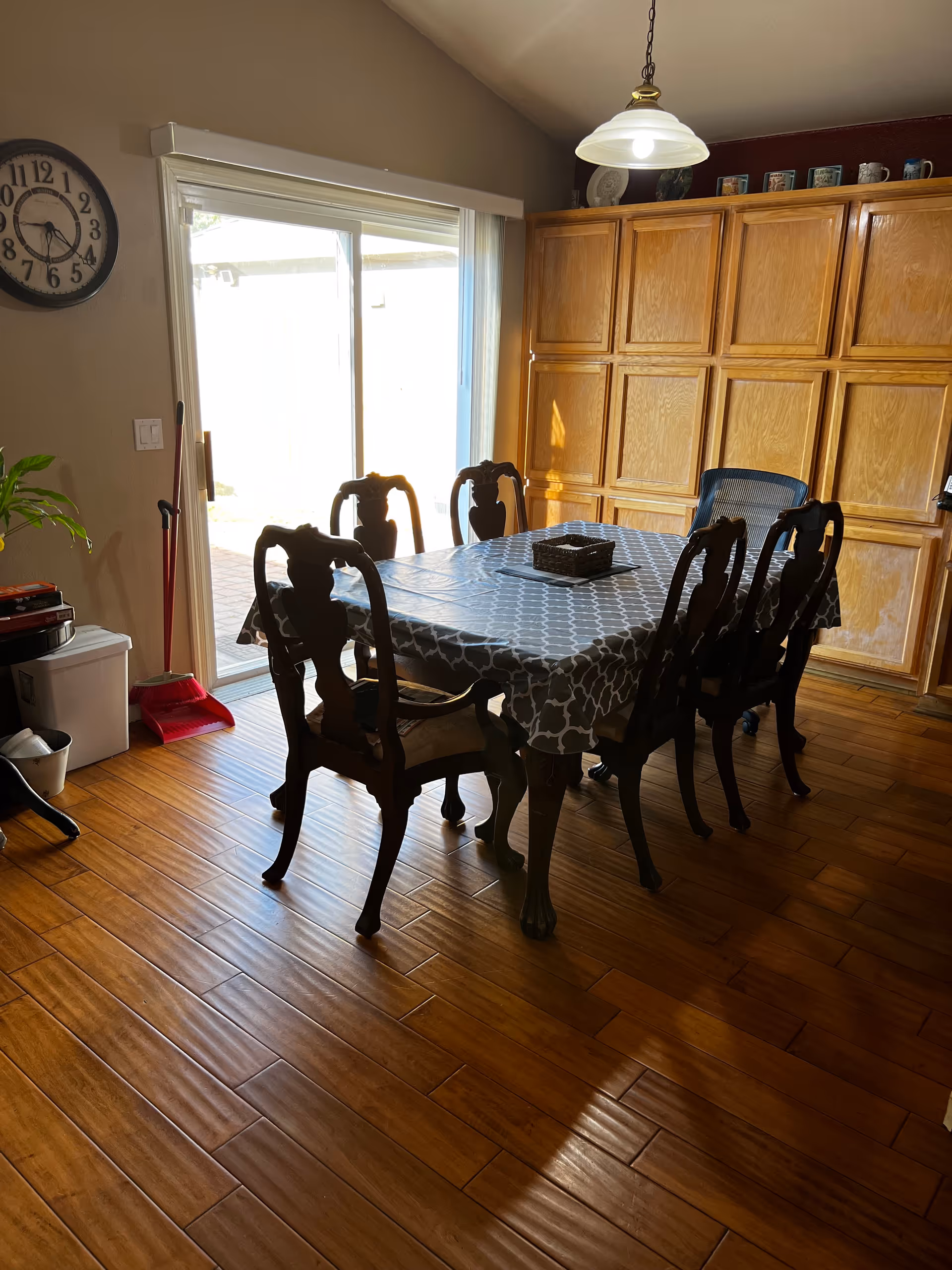 A dining room with a wooden floor, a rectangular table covered with a patterned tablecloth, and six chairs around it. There is a large wooden cabinet against the wall, a hanging light fixture above the table, a wall clock, and a sliding glass door letting in natural light.
