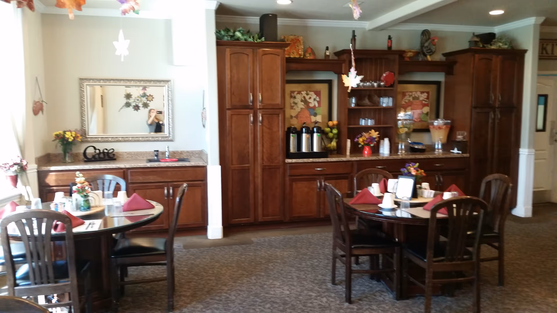 A dining area with two round tables set with maroon napkins, cups, and small flower arrangements. Behind the tables is a wooden cabinet and countertop with coffee dispensers, pitchers of infused water, and decorative items. A large mirror hangs on the wall above a smaller counter with flowers and a decorative 'Cafe' sign. The room has carpeted flooring and soft lighting.