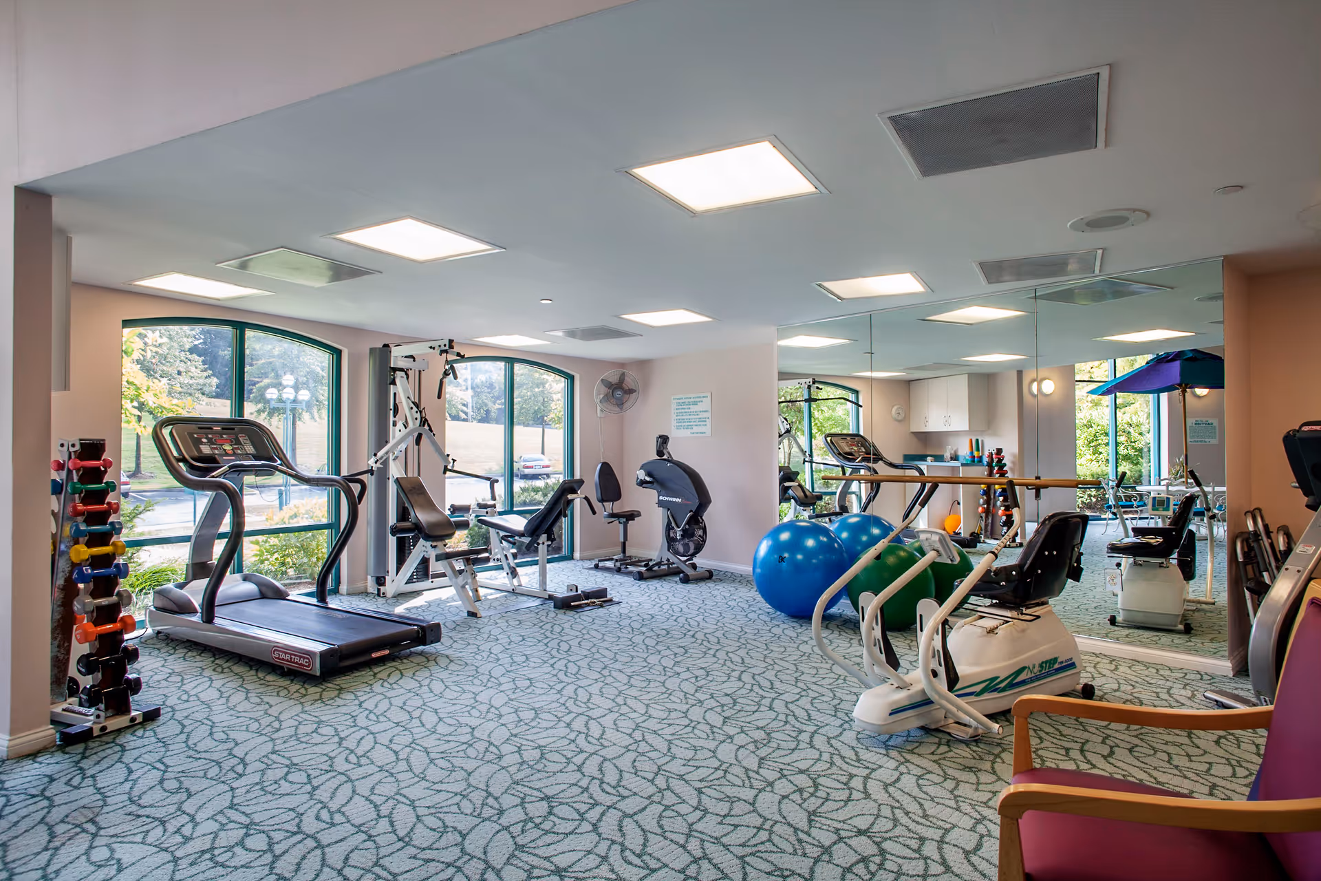 A well-lit fitness room with exercise equipment including a treadmill, stationary bike, weight machine, dumbbells, and large exercise balls. The room has large windows with views of greenery outside and a mirrored wall reflecting the equipment. The floor is carpeted with a patterned design, and there is a purple chair in the foreground.