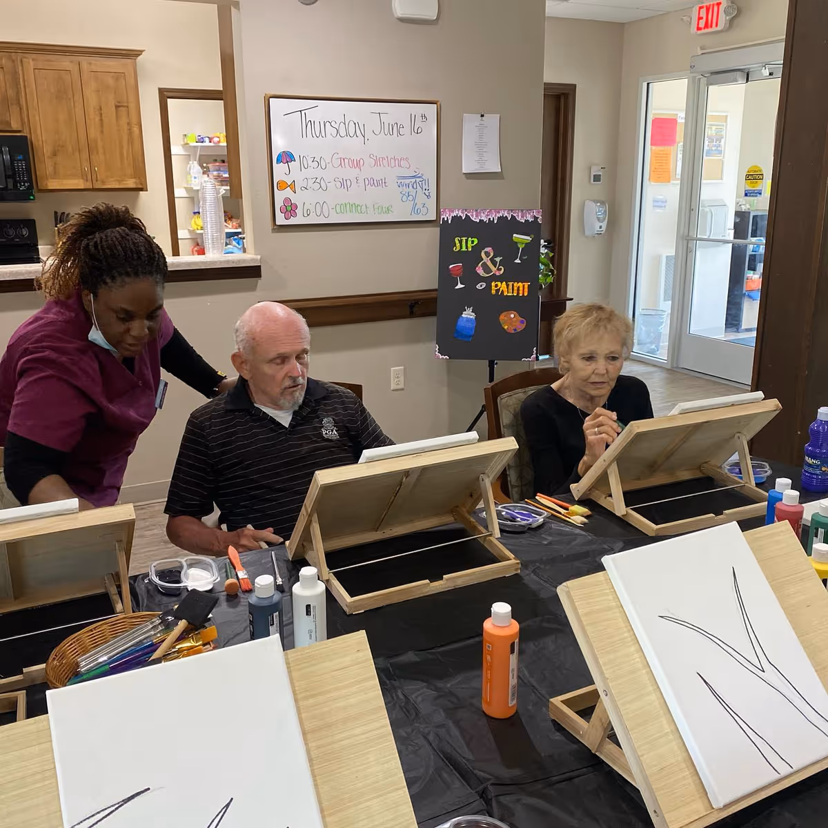Two elderly individuals seated at a table with easels and painting supplies, engaged in a painting activity. A caregiver in a purple uniform is assisting one of the elderly men. Behind them, a whiteboard displays the day's schedule including group stretches, sip and paint, and connect hour. A colorful sign reads 'SIP & PAINT'. The setting appears to be a common activity room in an assisted living facility.