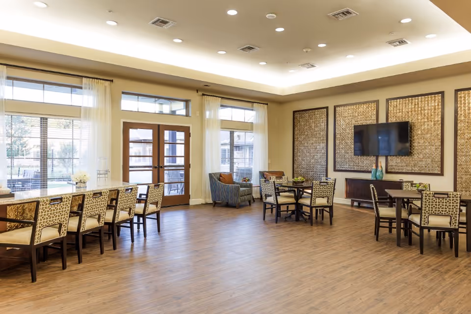A spacious and well-lit common area in a senior living facility with wooden flooring, multiple tables and chairs, two armchairs near large windows with sheer curtains, a wall-mounted flat-screen TV, and decorative wall panels. French doors lead to an outdoor area.