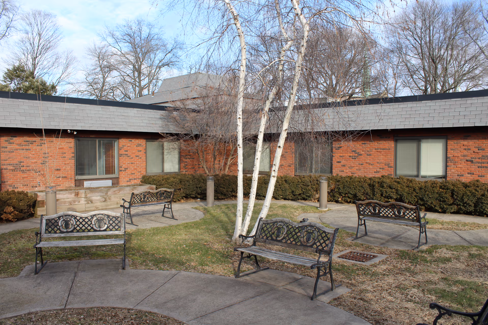 Outdoor courtyard area with several metal benches arranged on a concrete pathway surrounded by grass and leafless trees. The background shows a single-story brick building with windows and a gray shingled roof under a partly cloudy sky.