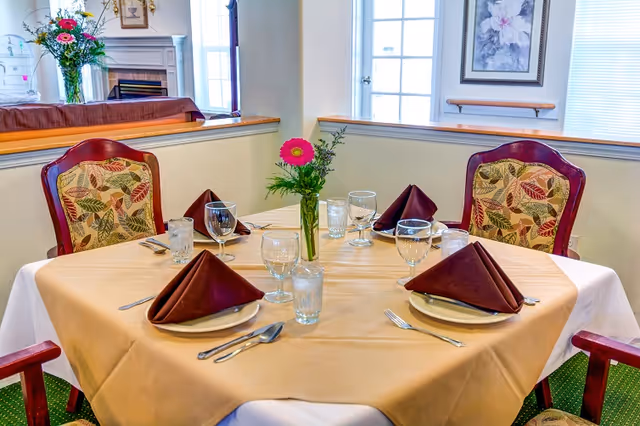 A dining table set for four with beige tablecloth and brown folded napkins on white plates. Each place setting has a fork, knife, spoon, water glass, and wine glass. A small vase with a pink flower and greenery is in the center. The background shows a cozy room with floral upholstered chairs, a fireplace, and framed artwork on the walls.