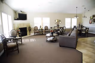 A spacious living room area with beige walls and carpeted floor. The room features a fireplace with a TV mounted above it, several chairs arranged around the room, a sofa, and two dining tables with chairs. Large windows allow natural light to fill the space.