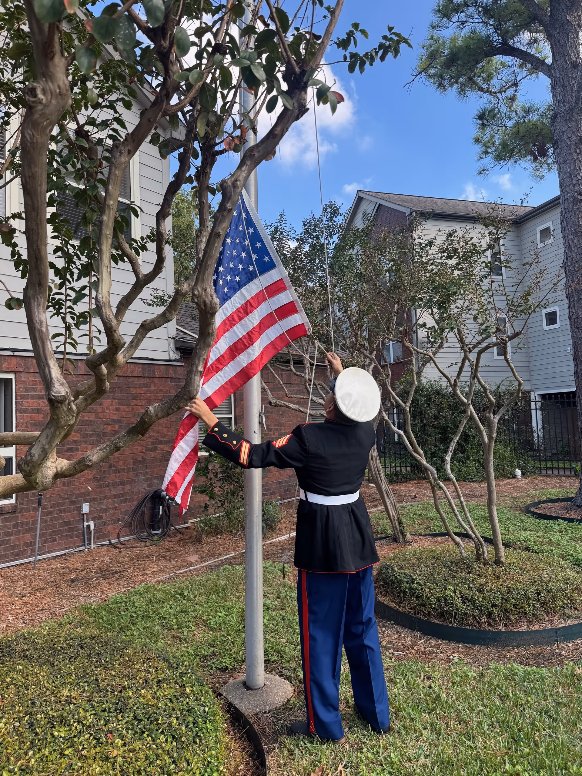 A person dressed in a formal military uniform is raising an American flag on a flagpole outside near a building with brick and siding exterior. The area has green grass, trimmed bushes, and several trees under a partly cloudy blue sky.