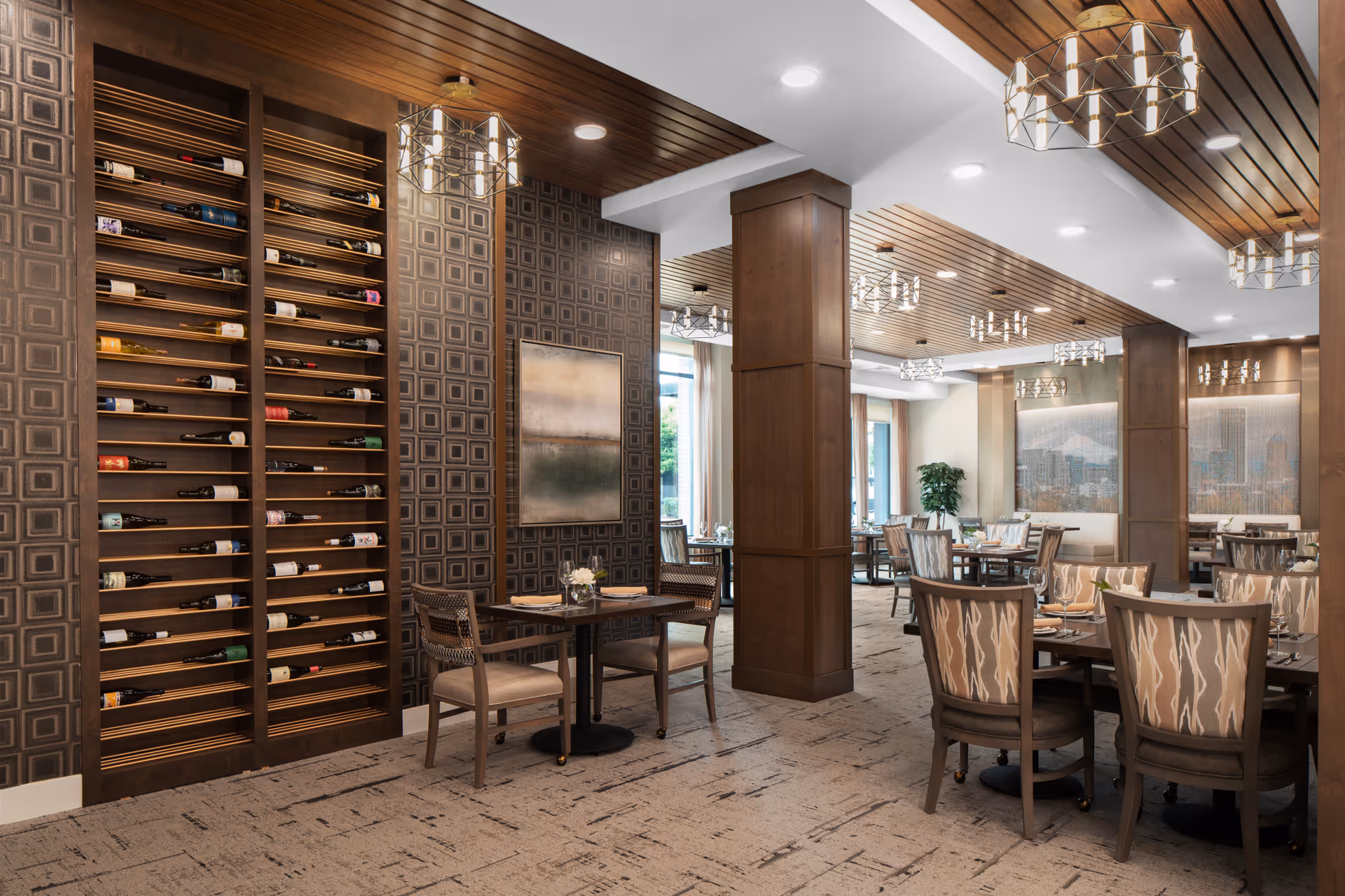 A spacious and elegant dining room with multiple tables and chairs arranged neatly. The room features a wooden ceiling with modern light fixtures, patterned wallpaper, and a large wine rack filled with bottles. There are large windows allowing natural light to brighten the space, and decorative artwork on the walls.