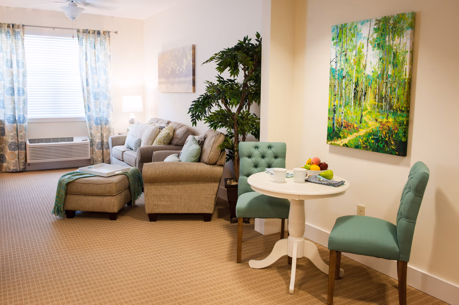 A cozy living room area with a beige sofa, matching ottoman, and armchair. There is a small white round table with two green upholstered chairs. The room has beige carpet, a large window with floral curtains, a potted plant, and two paintings on the walls.