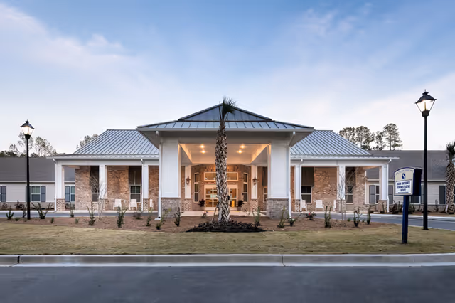Front exterior view of Sprenger Health Care Bluffton building with a covered entrance, brick and siding facade, two lamp posts, and a sign indicating main entrance and administrative office.