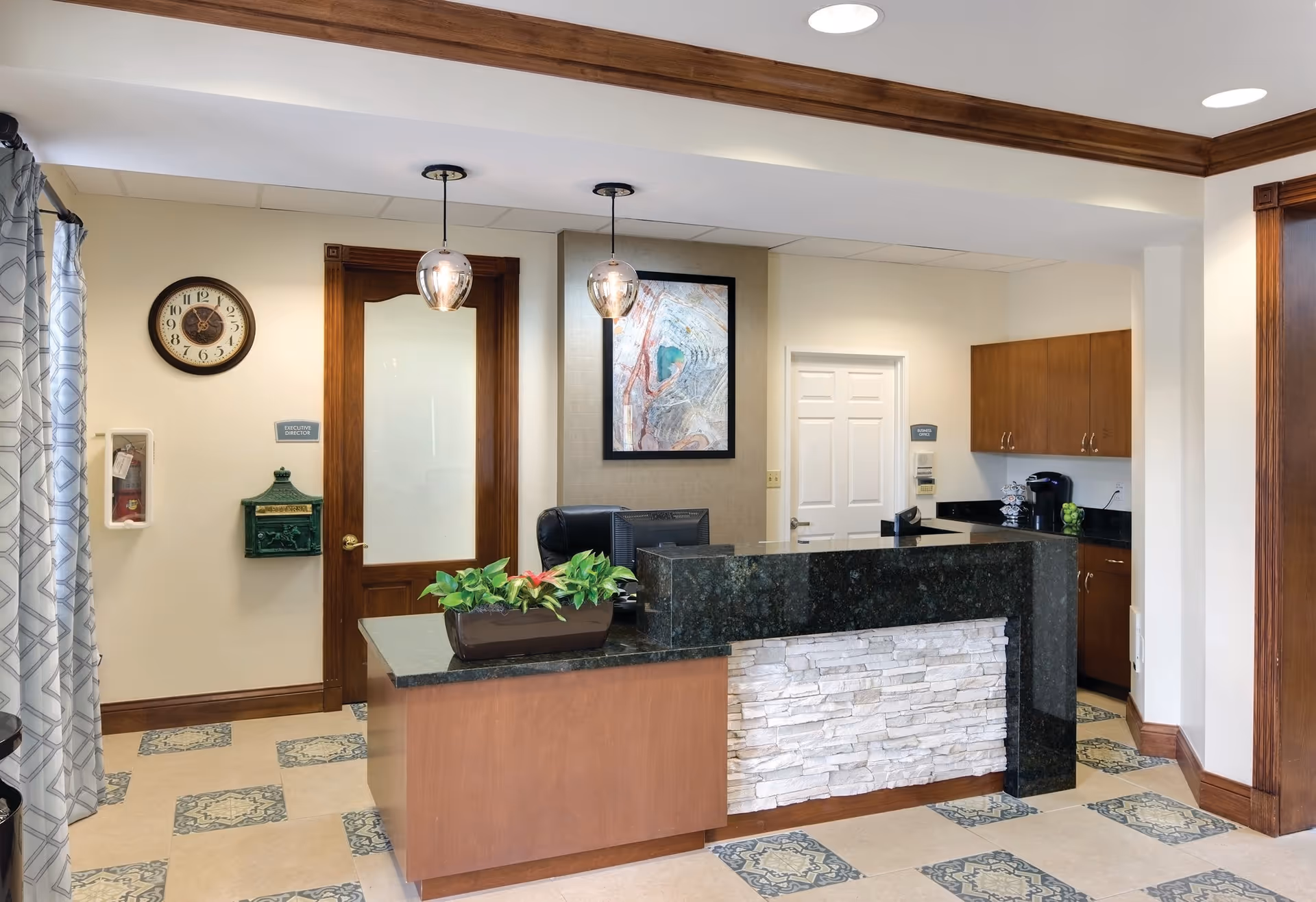 Reception area with a dark granite countertop desk featuring a stone facade, a potted plant on the desk, two hanging pendant lights, a wall clock, a wooden door labeled Executive Director, a white door labeled Business Office, wooden cabinets, a coffee maker, and a framed abstract artwork on the wall.