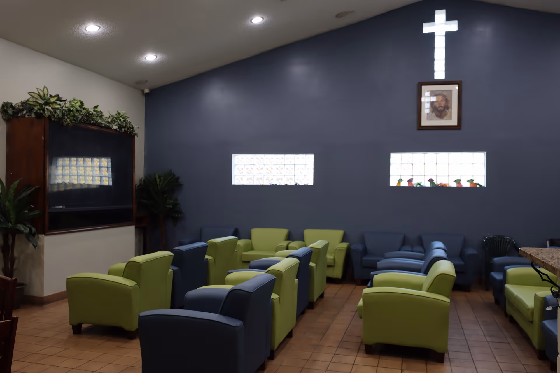 Communal lounge with rows of green and blue armchairs, glass-block windows, a mounted TV, and an illuminated cross on the wall.