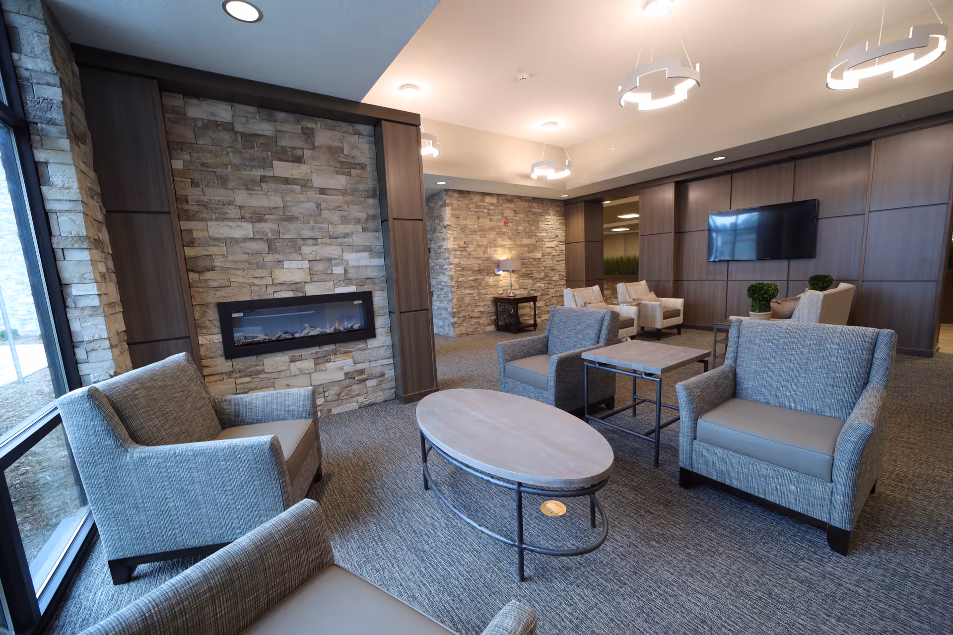 Bright, modern lounge with upholstered armchairs arranged around an oval coffee table, a stone fireplace, and a wall-mounted TV.