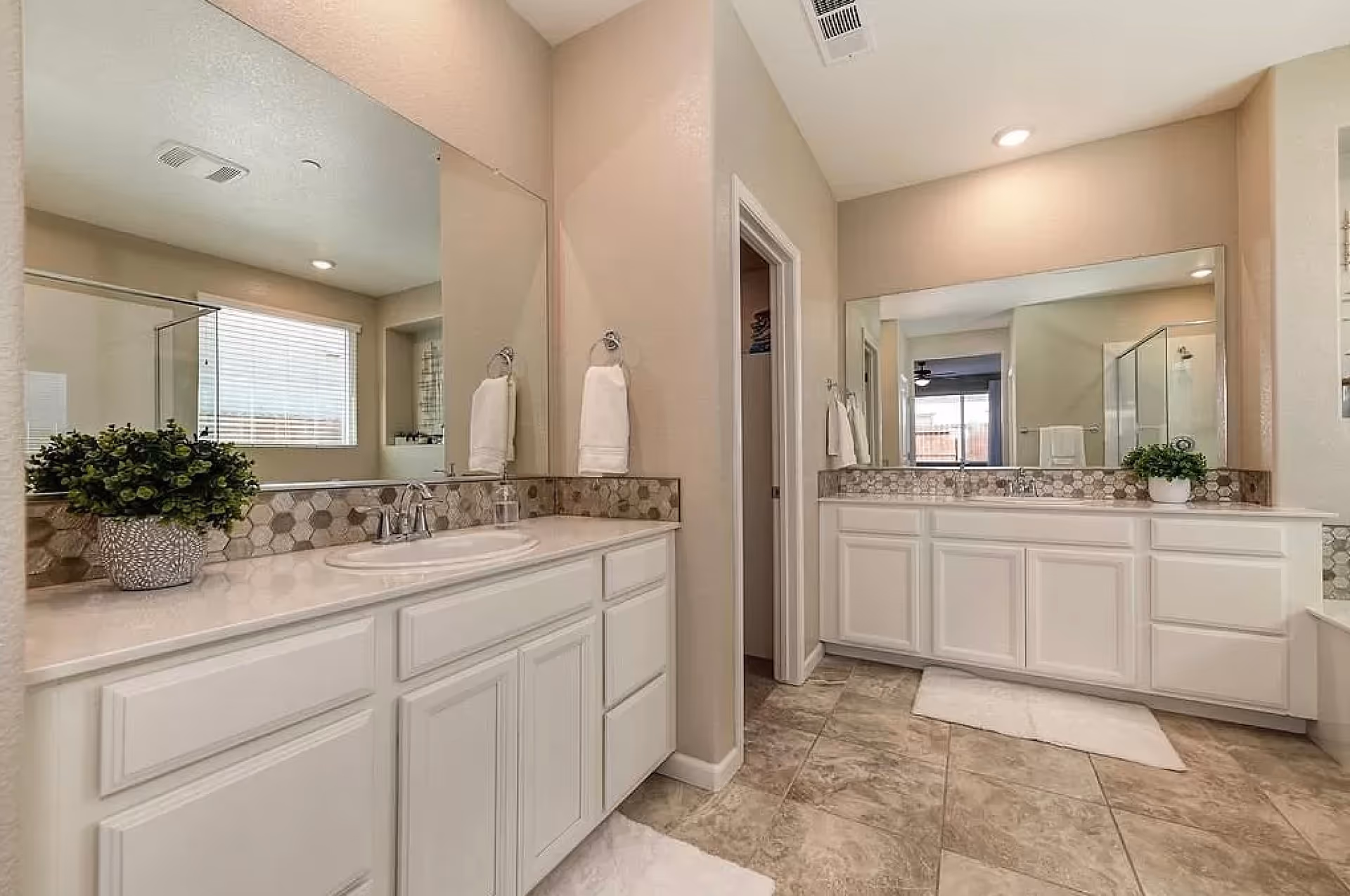 A spacious bathroom with two large mirrors above white cabinets and sinks. The room features beige walls, tiled floors, a window with blinds, and decorative plants on the countertops. There are white towels hanging on towel rings next to each sink.