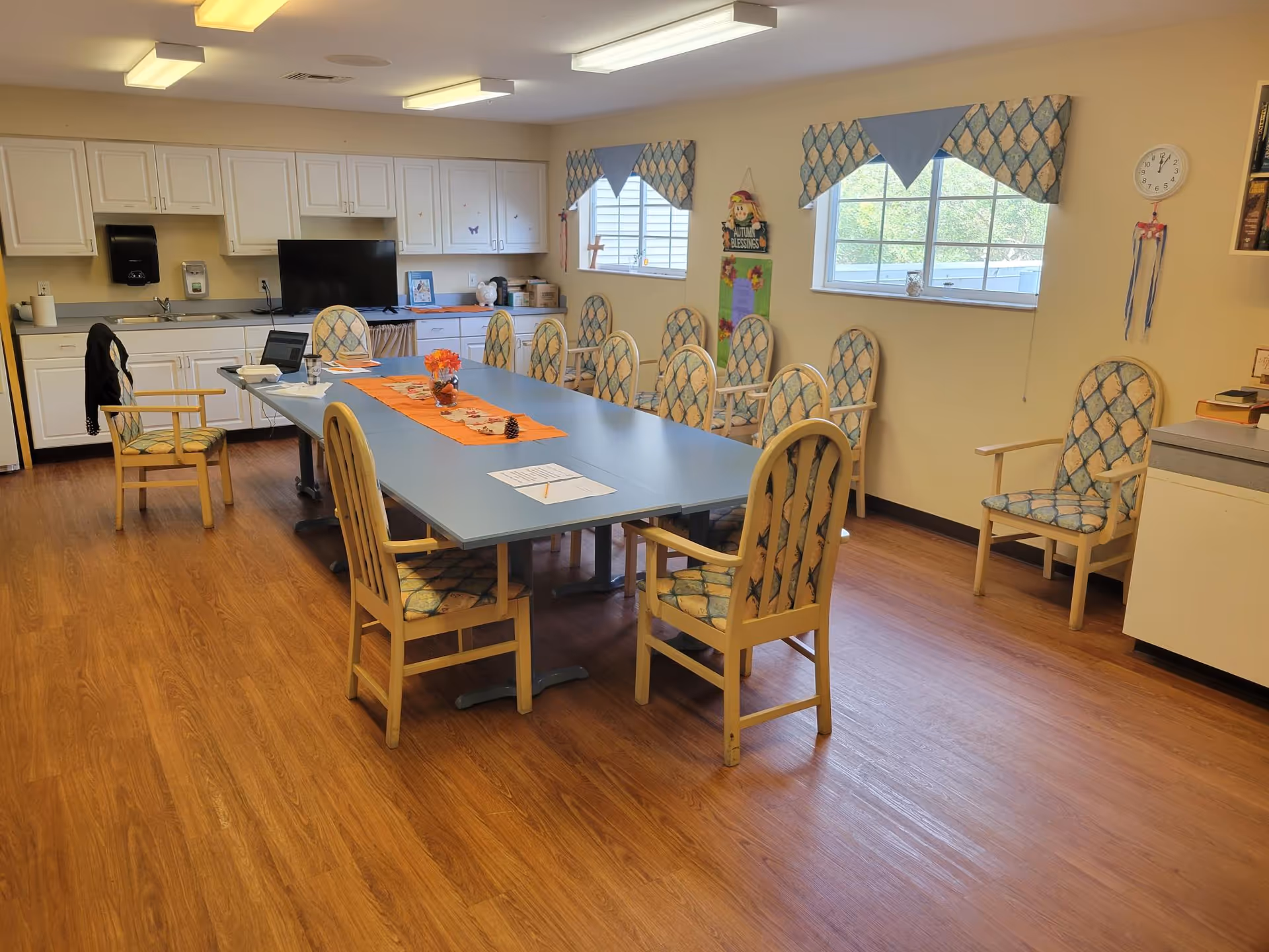 A well-lit room with a long rectangular table surrounded by cushioned chairs with patterned upholstery. The table has an orange runner with decorative items including a small vase with flowers and pine cones. The room has wooden flooring, white cabinets along one wall, two windows with patterned valances, and a clock on the wall. There is a laptop and some papers on the table.
