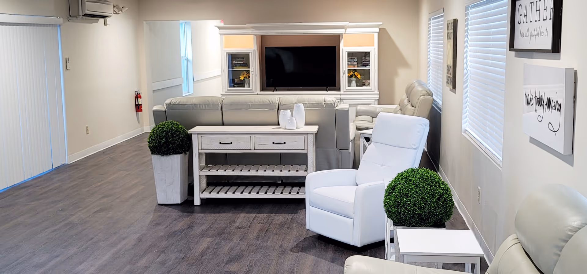 A bright and spacious living room area with light-colored leather sofas and armchairs arranged around a white wooden console table. A large flat-screen TV is mounted on a white entertainment center against the far wall. Two decorative green plants in white pots flank the console table. The room has wood flooring, light-colored walls, and windows with white blinds. Inspirational wall art is visible on the right wall.