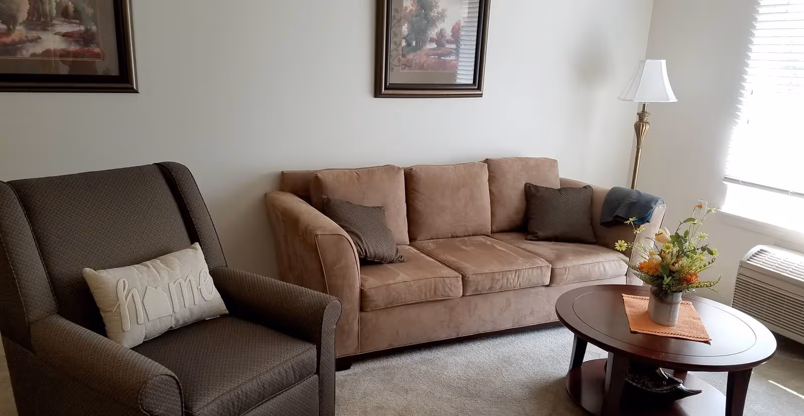 A cozy living room with a brown upholstered sofa adorned with two dark pillows, a dark patterned armchair with a decorative pillow that says 'home', a round wooden coffee table with a flower arrangement on an orange placemat, a floor lamp, and a window with blinds letting in natural light.