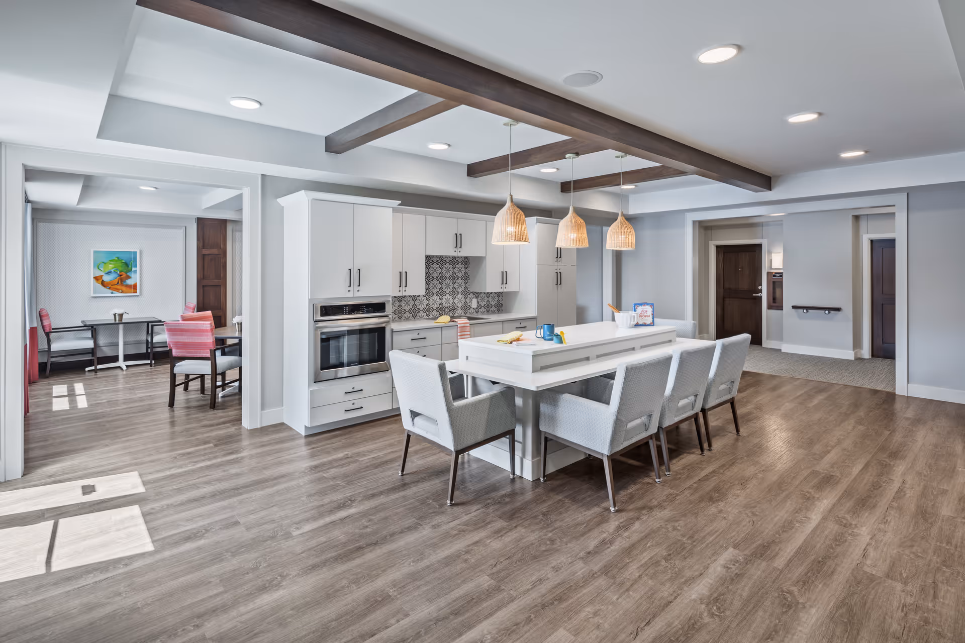 A modern communal kitchen and dining area with white cabinetry, built-in oven, and a central island with seating for six. The space features wood flooring, recessed lighting, and three hanging wicker pendant lights. Adjacent to the kitchen is a dining area with tables and chairs, and the walls are painted light gray with decorative wooden ceiling beams.