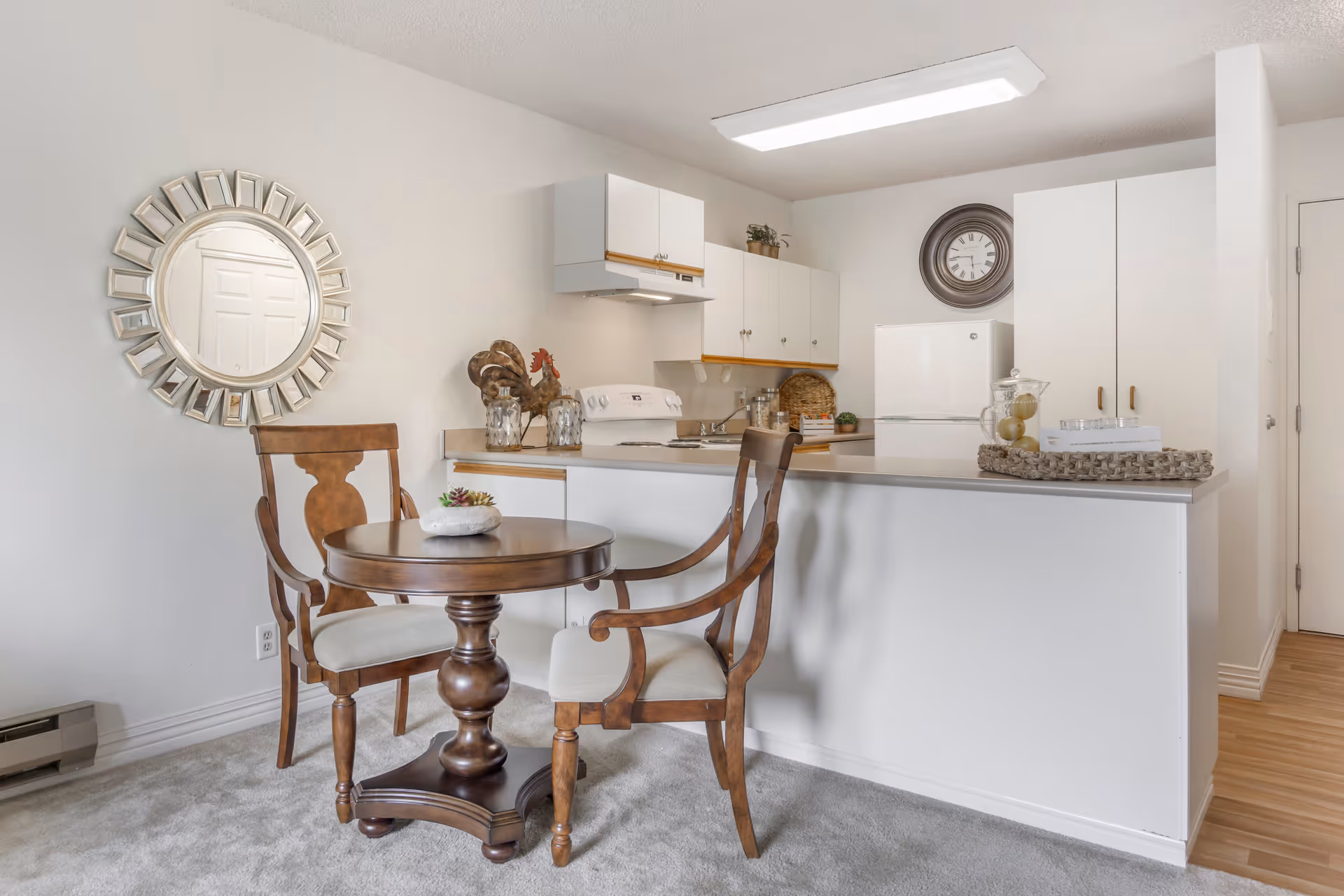 A small kitchen and dining area in a senior living facility. The kitchen features white cabinets, a white stove, and a white refrigerator. A round wooden table with two wooden chairs with cushioned seats is placed in front of the kitchen counter. A decorative round mirror hangs on the wall to the left, and a large wall clock is mounted above the refrigerator. The floor is carpeted near the dining area and transitions to wood flooring in the kitchen.
