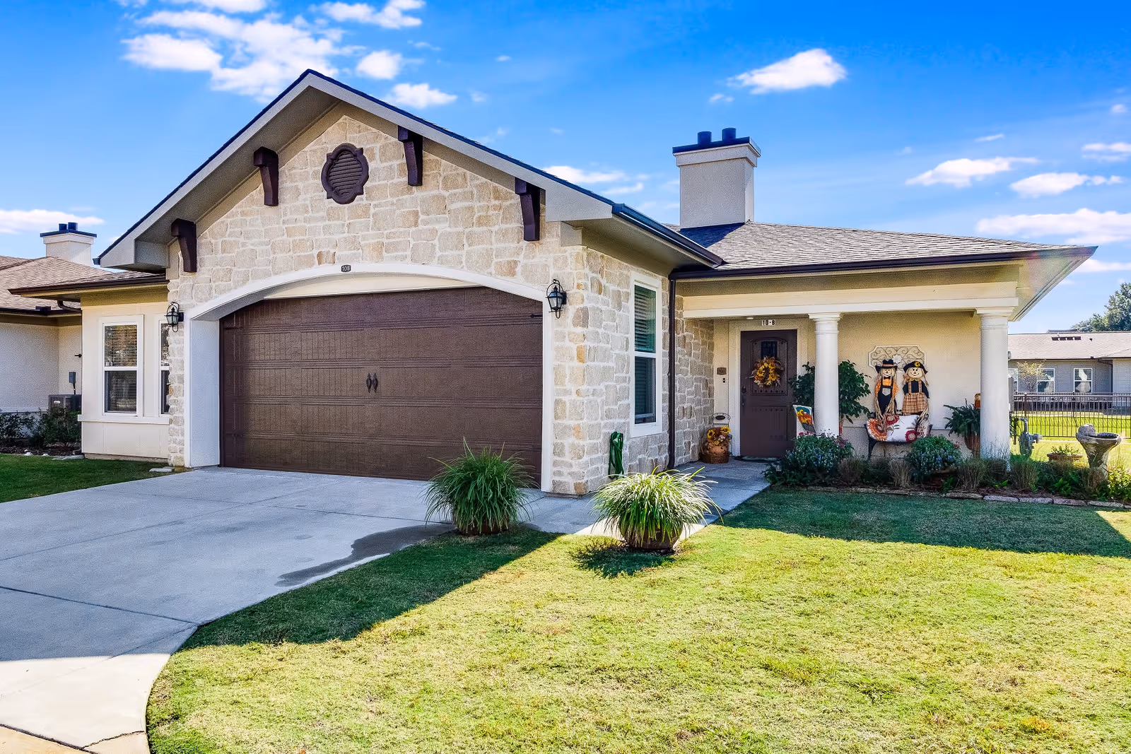 Single-story stone-faced house with a two-car garage, front porch, and manicured lawn under a blue sky.