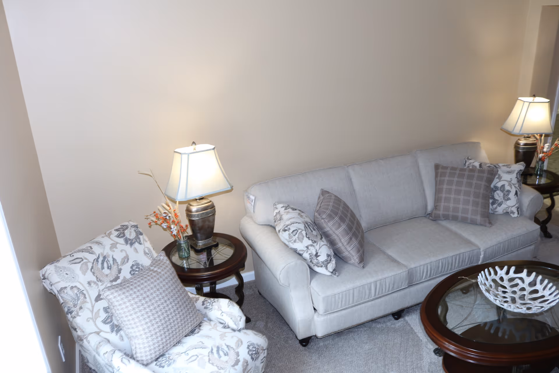 A cozy living room area featuring a light gray sofa with patterned throw pillows, a matching armchair with a houndstooth pillow, two wooden side tables each with a lamp and decorative items, and a round glass coffee table with a white coral-like centerpiece. The walls are painted beige and the floor is carpeted.