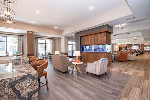 Spacious senior living facility common area with modern decor including a large built-in aquarium, comfortable seating with armchairs and sofas, a granite countertop with bar stools, and large windows letting in natural light.